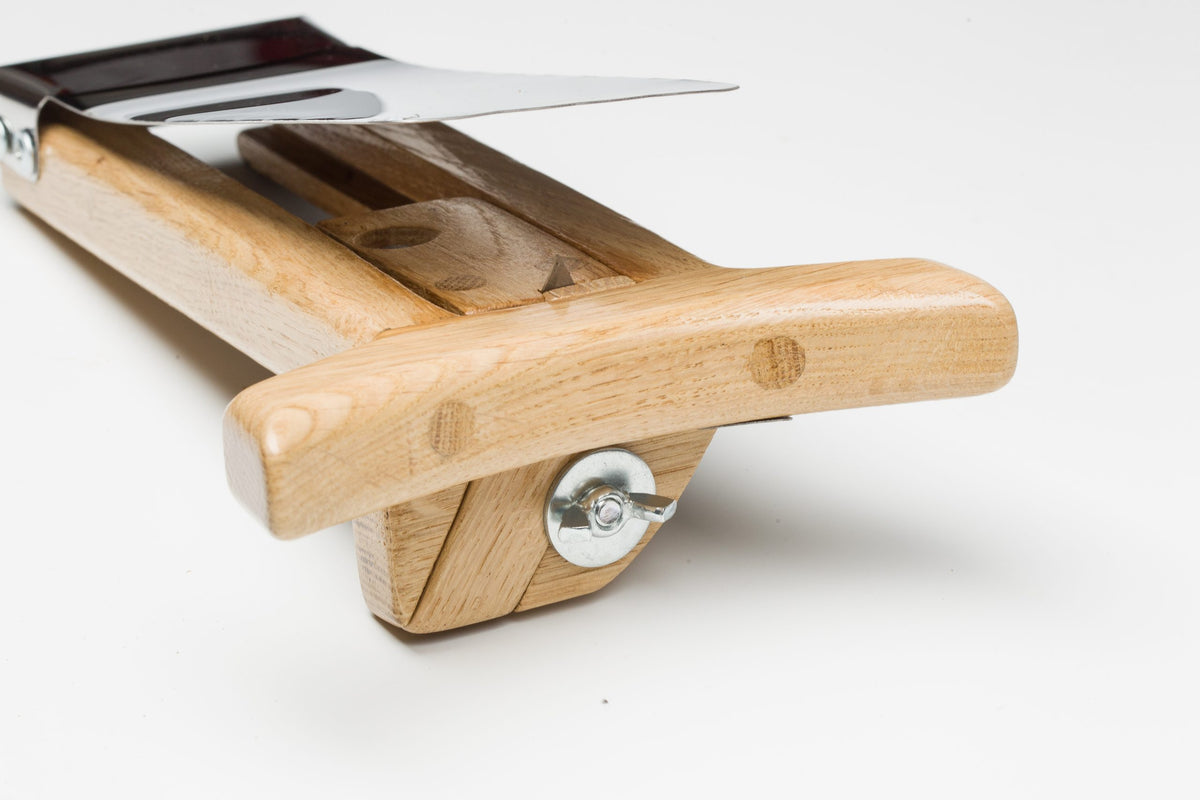Low-angle close-up of the wooden hat brim cutter showing the curved handle, adjustable stop, wing-nut lock, and angled metal blade used for clean, controlled brim trimming.